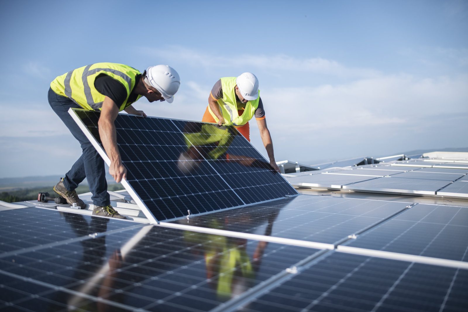 Two installers in hard hats and safety vests lifting a solar panel into place